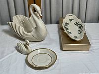 Four Lenox porcelain items displayed on white cloth, including a large swan-shaped candy dish, small swan figurine, gold trim saucer, and a Holly pattern candy dish on a box.