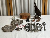 Full lot containing wooden box, bird figurine, metal trivets, small metal teapot trinket, ceramic stand, spoon and miniatures on white tablecloth with curtain background.