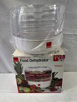 Front view of Ronco 5-Tray Food Dehydrator in shrink wrap atop original box showing product image with fruits and vegetables down front.