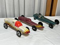 Three vintage wooden derby style toy cars showing front and side views, all with flame designs and colored cockpit covers.