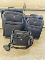 Front view of large, medium, and small black SwissGear soft shell luggage pieces showing their front zipped compartments and logo.