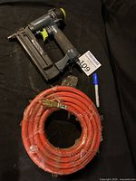 Photo of the Pro-Pulse 18 gauge brad nailer and coiled red 3/8 air hose on black backdrop with MaxSold auction tag and marker pen for scale.