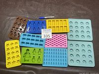 Photo showing eleven assorted chocolate or ice molds in various shapes and colors including stars, hearts, robots, gingerbread men, and round balls, with a Sharpie pen for scale and a MaxSold lot number card.