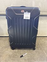 Closed black hard shell suitcase with four wheels and a top handle, placed on concrete floor with a marker for scale.