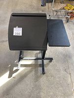 Front view of black adjustable laptop table with curved lip on tilted surface and adjacent flat side shelf on caster base.