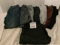 Seven pairs of men's pants and one pair of men's shorts displayed folded on a bed in various colors including blue denim, gray, black, brown, and rust shorts.