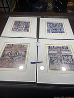 Four framed Paris style art prints laid flat on a surface, showing detailed Parisian storefronts and street scenes in watercolor style.