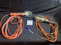 Overview of two handheld trouble lights with protective cages and long extension cords, one orange handle and one black handle NOMA brand.