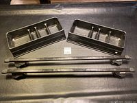Four black adjustable roof rack cross bars laid out horizontally with two black plastic trunk caddies above them on a black surface.