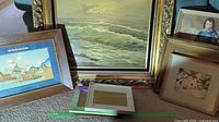 Wide view showing large seascape painting in ornate gold frame, smaller framed maritime scene, and other stacked framed pieces.