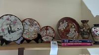 Photo showing five decorative Asian-themed plates arranged on a shelf along with two books on Japanese ceramics and Imari Satsuma.