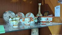 Front view of the Asian themed ceramic set with miniature jugs, cups, plates, and vase on glass shelf.