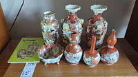Lot overview showing seven vases and bowl in detailed Asian Satsuma style with painted figures and patterns alongside the Satsuma guide book