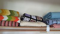 Full view of the six folded blankets stacked on a shelf showing a range of colors and textures including crocheted and woolen items.