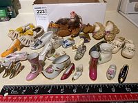 Wide view of the collection of miniature shoes in ceramic, pottery, glass, and other materials. Includes various floral decorated shoes and some gold trimmed ones arranged with a ruler for size reference.