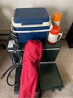 Overview photo showing large green cooler, smaller blue and white cooler, Coleman camp stove, two thermos containers (orange/white and solid orange), and red sports chair in bag on top.