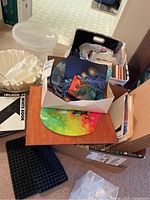 Photo showing artistic mouse pads with abstract and space designs, crafting books, white and black notebooks, plastic storage container, and assorted crafting papers and cards, displayed on and around cardboard boxes.