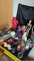 Photo showing assorted figurines including wooden tattooed dolls, plush doll, painted armadillo and other decorative items on a table against a black cloth background.