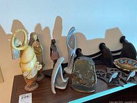 Multiple figurines including two ceramic lady figurines, carved tribal figure, brass bookends, modern sculptures and wooden shelves arranged on a shelf.