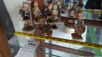 7 Hummel figurines arranged on glass shelf with measuring tape for size reference