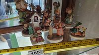Front view of 7 assorted Hummel figurines in pastel colors, showing various child characters with props such as books, animals, and a birdhouse. The figurines range in size up to about 6 inches tall. Measuring tape included for scale.