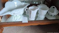 Full view of 5 milk glass items arranged on mirrored wooden surface including bowl, pitcher, sugar bowl, basket, and rectangular dish.