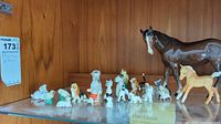 Wide view of various small porcelain animal figurines including two horses and multiple small dogs, cats, and birds arranged on a wooden shelf.