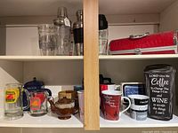 Shelf with bar shakers, bottle carrier, teapot, and various mugs.