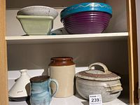 Overall lot showing five pottery items on two shelf levels: green bowl, purple and blue bowl, beige casserole with lid, tan jar, and blue-green glazed mug with animal scene.