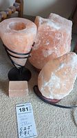 Three pink Himalayan salt items including two lamps and one candle holder on a carpeted floor with lot tag and measurement tape nearby.