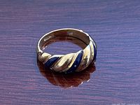 Front view of ring placed on wooden surface showing alternating polished gold and blue enamel twist design