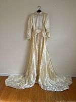 Full front view of cream satin wedding gown hanging on a wall, showing bodice, sleeves and cathedral-length train