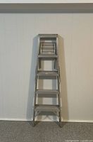Front view of the 5ft aluminum folding ladder fully closed and standing upright against a white paneled wall.