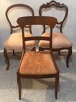 Photo showing three vintage wooden chairs with peach velvet upholstery from a front angle, highlighting different back designs and curved wooden legs.