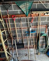 Photo showing various wooden and metal handled garden tools including shovels, rakes, broom, and a green garden hose hung on wall