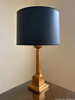 Full front view of the gold lamp with black drum shade on a wood surface, showing overall condition and proportions.