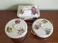 Front view of three white porcelain floral casserole dishes including one rectangular, one round, and one oval with gold handles on a wooden table.
