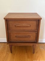Frontal view of the mid century teak end table nightstand with two drawers on a hardwood floor against a white wall.