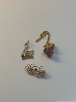 Three jewelry items on white background: Virgo pendant, gold brooch with blue stone, and tie tack with purple stone.