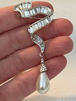 Brooch held in hand showing the scale and detail of the rectangular rhinestone arrangement and dangling faux pearl teardrop.