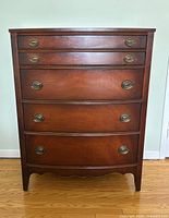 Front view of vintage mahogany highboy dresser showing five drawers with brass oval handles and curved front panels.