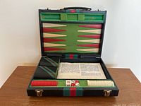 Open black travel backgammon case showing triangular pattern backgammon board interior in green felt with red and white points, two striped dice cups, red and white checkers, dice, and instruction sheet.