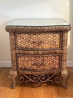 Front view of woven wicker and rattan night table showing two woven drawers with rattan handles, and wrought iron scrollwork detail on the base and legs beneath a glass top.