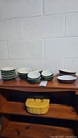 18 pieces of Hall stoneware dishes arranged on a wooden shelf against a concrete wall. Includes various bowls and plates in brown and green stoneware.