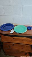 Three ceramic plates displayed on a wooden surface including two 9.5 inch plates (blue and aqua green) and one small round blue item.