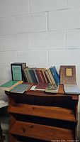 Photo showing 36 vintage books arranged on a wooden shelf against a white brick wall. Includes various sizes, colors, and conditions, one with a wooden cover titled 'Rules of this Tavern'.