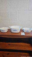 Front view of three CorningWare Blue Cornflower casserole dishes of different sizes arranged on wooden surface against white brick wall