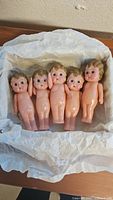 Five vintage plastic dolls approximately 4 inches tall arranged in a box with tissue paper. Dolls have painted hair and distinct facial features.