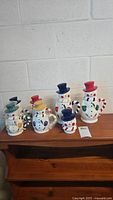 Full set of six Temptations snowmen mugs displayed on wooden shelf, showing all with hats and colorful details.