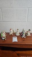 Seven Christmas-themed ornaments including Santa and snowmen arranged on wooden surface against white brick wall.
